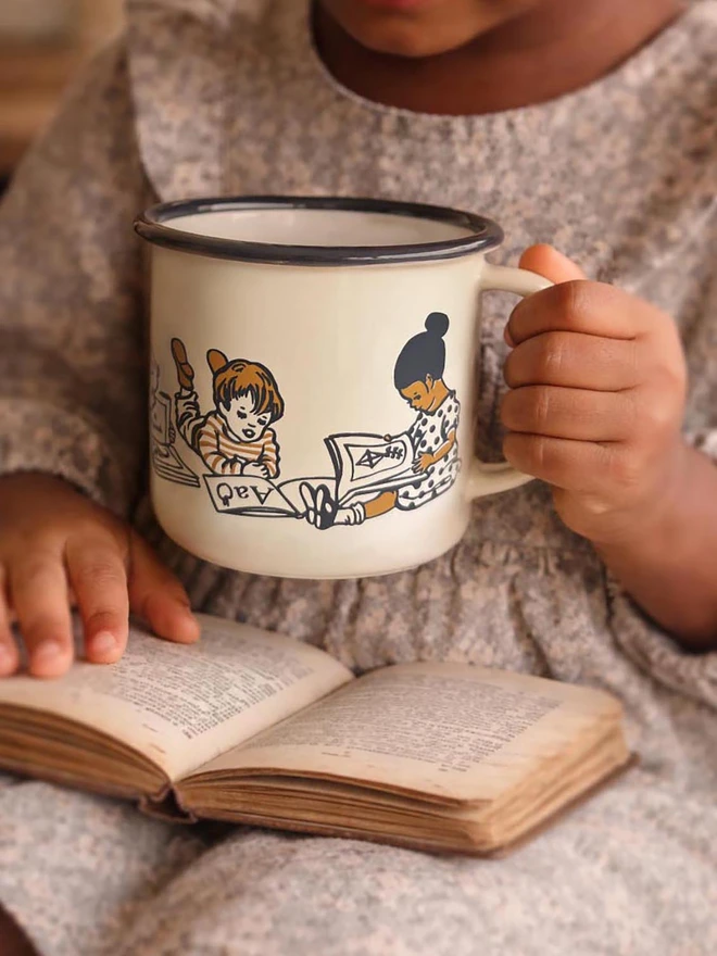A vintage style enamel mug featuring a traditional vintage style illustration of children drawn in a vintage story book style reading books cream in colour with accents of ochre brown and grey