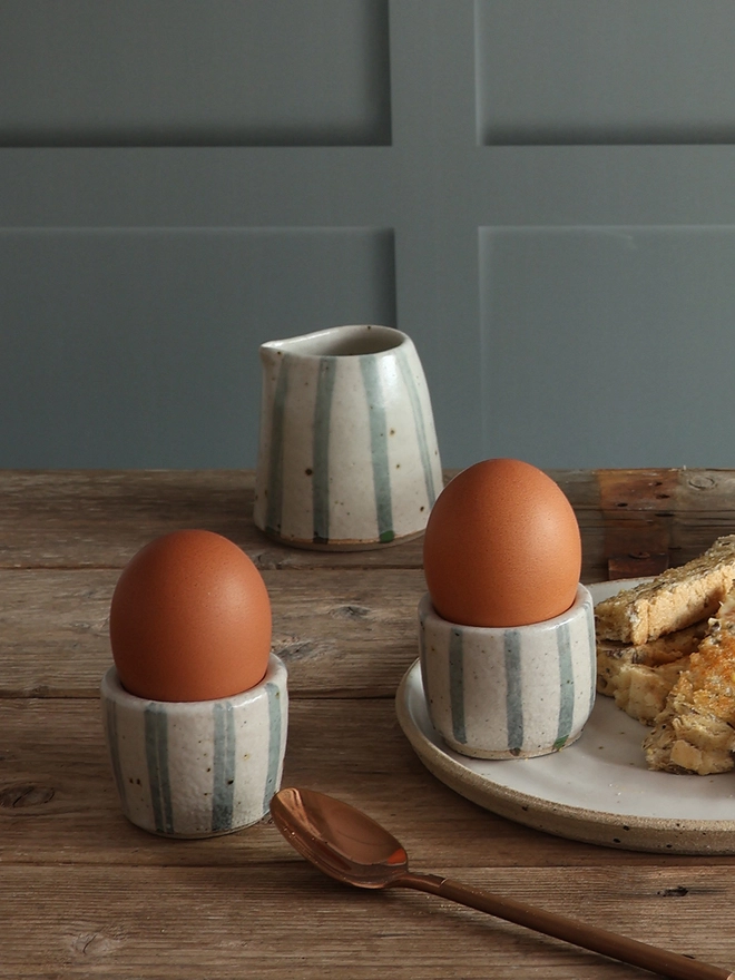 Two Handmade Ceramic Stoneware Egg Cups in Blue and White Stripe holding boiled eggs sit on a wooden surface next to a brass spoon and a plate of toast. A small jug in the same blue and white stripe design sits in the background. 