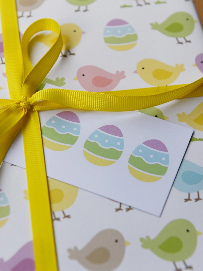 A gift wrapped in pastel spring chicks wrapping paper tied with yellow ribbon has a gift tag with three easter eggs on it.