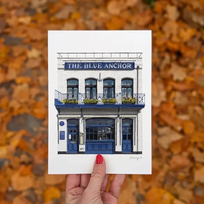 Hammersmith The Blue Anchor Giclée Print (Unframed), a woman holding the print of a blue pub against a colourful backdrop. 