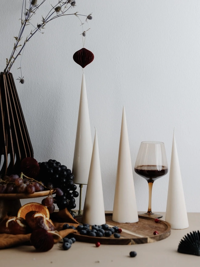 White Christmas Tree Cone Candles (Set Of 3). Four cream cone candles of different heights on a wooden tray, styled with grapes, blueberries, a glass of red wine and a burgundy paper ornament. The modern sculptural shapes suit seasonal entertaining and minimalist interiors.