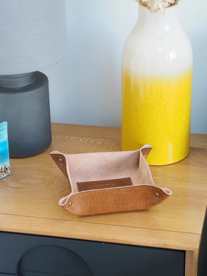 Personalised message leather coin tray. Hand dyed tan brown leather tray with a square stitched to the base which is personalised with a short but sweet message. The tray sits on a side table next to a lamp and vase.