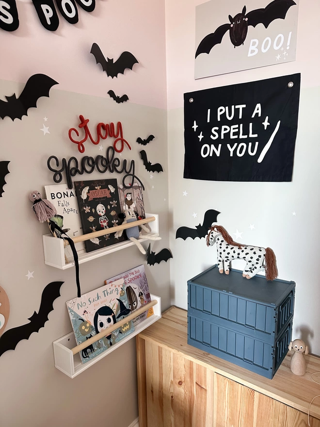 'stay spooky' knitted wire word sign hanging above some bookshelves in a child's reading corner or book nook surrounded by Halloween prints and decorations for the wall. 