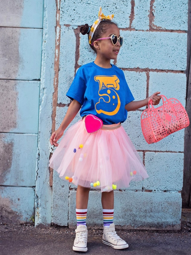 Girl wearing a blue t-shirt and peach pom pom tutu with neon pom poms, standing in front of a wall and holding a bag