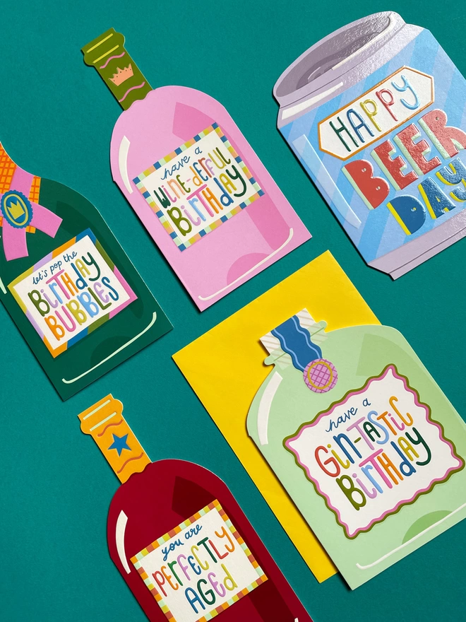 A flat lay of colourful birthday cards from the Raspberry Blossom ‘Cheers’ greeting card collection. Cards include die cut shapes of gin, beer, wine and champagne bottles all with a playful pun-filled birthday message