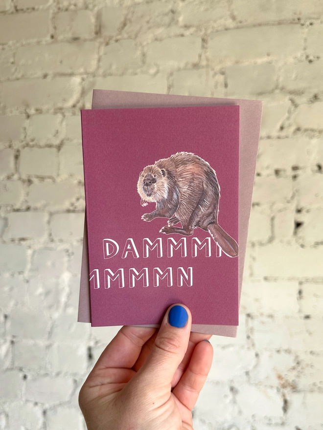 a pinky purple greetings card featuring an illustration of a beaver with the text “dammmmmn” with a muted purple envelope