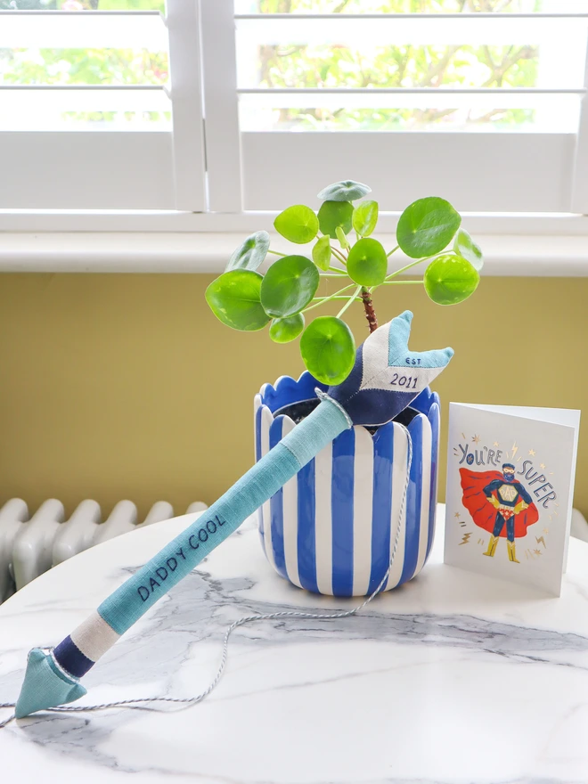 A handmade linen arrow wall hanging in shades of blue and navy, embroidered with the words “DADDY COOL” along the shaft and “2011” on the fletching. A white and grey cord is attached for hanging.