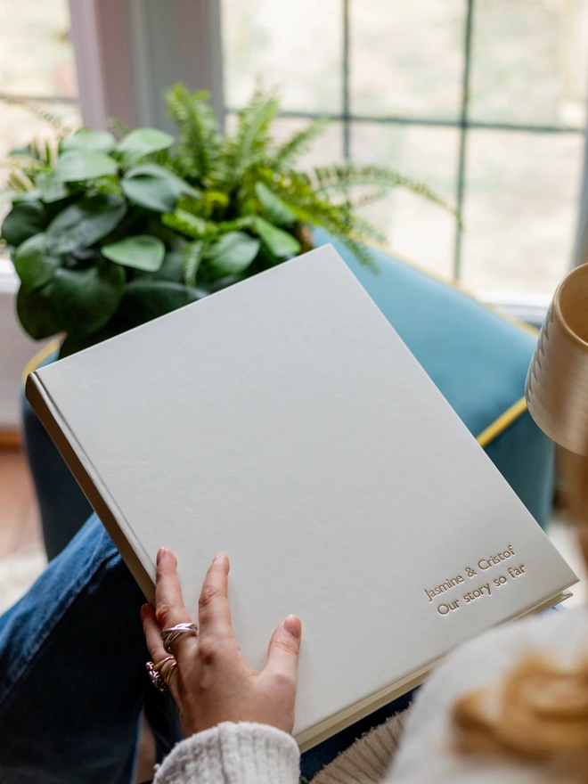 A woman is sitting in a chair holding a white A4 sized photo album. The album has been printed on the front in rose gold writing 