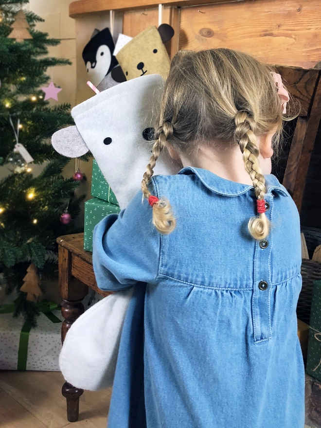 A young girl wearing a denim dress stands in front of a Christmas tree hugging a handmade felt polar bear stocking.