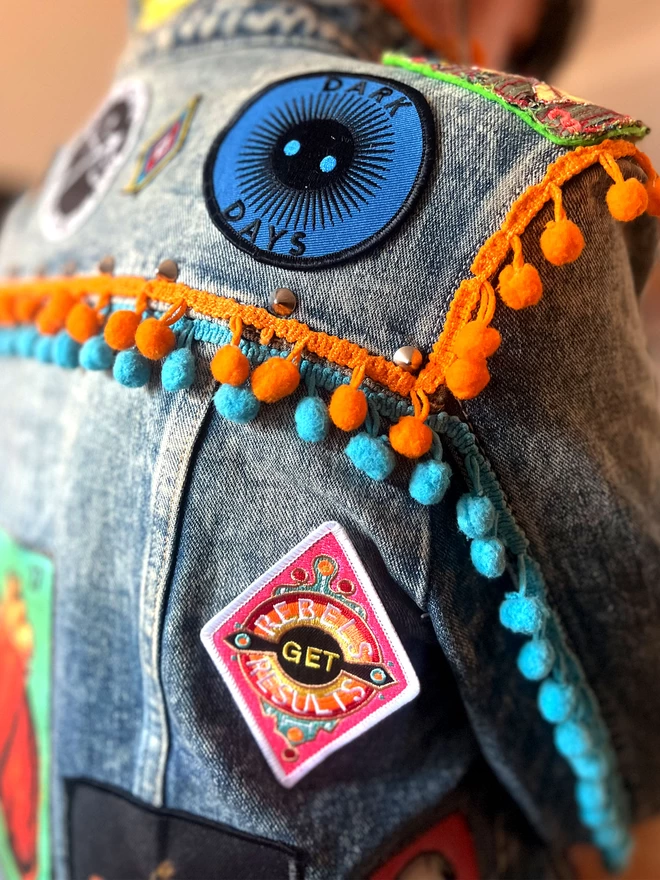 The patch is on a denim jacket with blue and orange trim detail and other patches. The patch is a diamond shape embroidered patch with “Rebels Get Results” at the centre. The design features pink, white, red, yellow and black colours and has a white border.