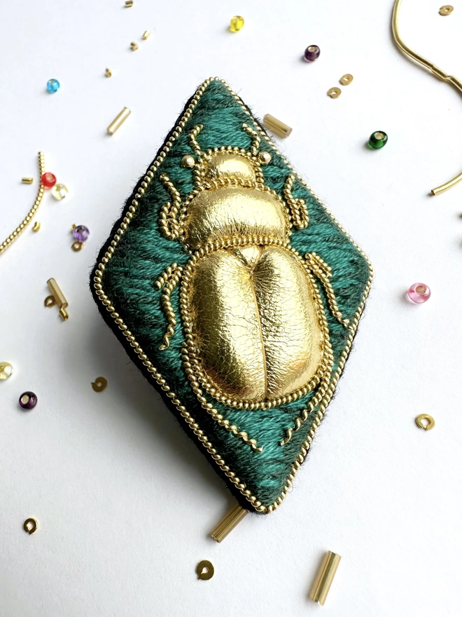 Diamond shaped brooch with golden beetle on a green background with beads and gold wire in the background 