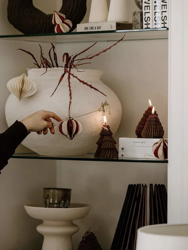 Brown Christmas Tree Warm Candles (Set Of 2). Glass shelves styled with lit chocolate brown Christmas tree candles, cream ribbed totem candles and swirled pillars, alongside a textured white vase, paper baubles and stacked books. The sculptural candles add warmth and a modern festive touch to a styled alcove.