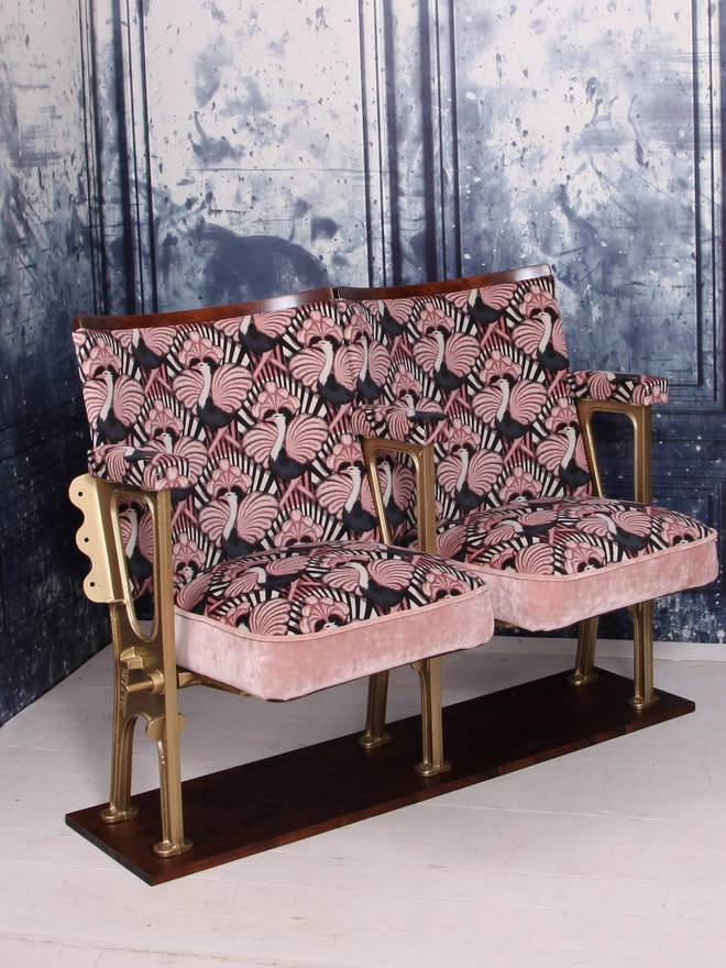 Set of two vintage cinema seats upholstered in a pink ostrich Art Deco design velvet with pink panel and piping against a blue marbled wall.  Seats are down