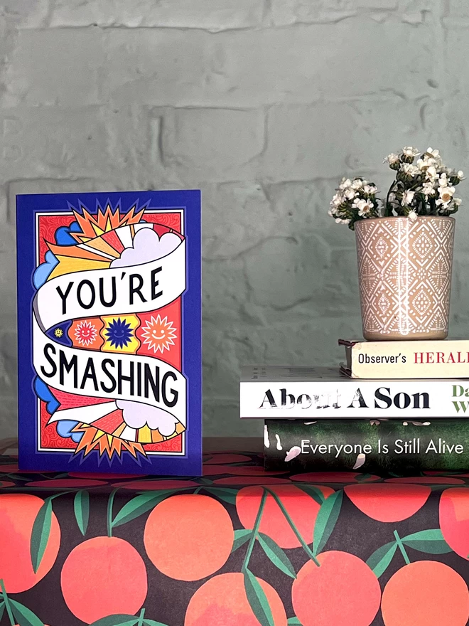 A blue greetings card featuring “You’re Smashing” written in black on a white swirl across a vibrant design of the sky featuring clouds and drawings of the sun with smiling faces, sits on a shelf covered with a wrapping paper with an illustration of oranges. Next to the card is a small white plant in a pink pot plant, on top of three books.