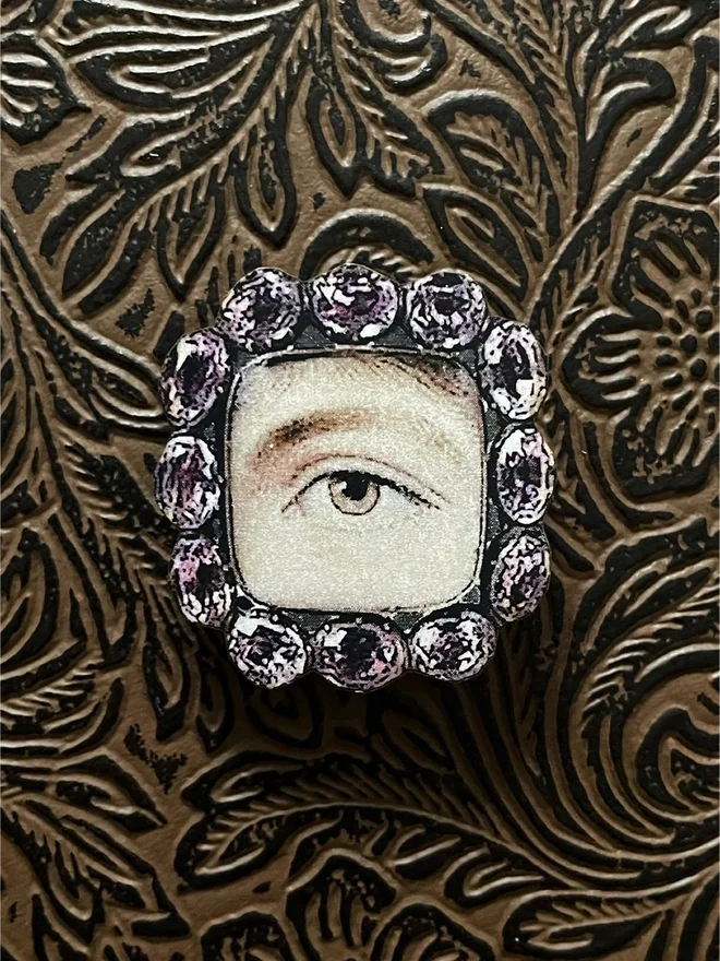 a lovers eye brooch, eye painting is surrounded by light purple crystals.  The brooch is a printout on wood