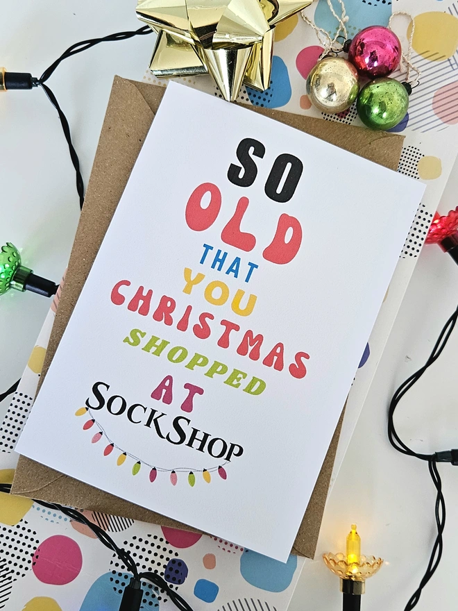 A photograph of the front of a humorous A6 greeting card illustrated using brightly coloured text and the retro shop logo. The text says so old you Christmas shopped at Sock Shop with string christmas lights