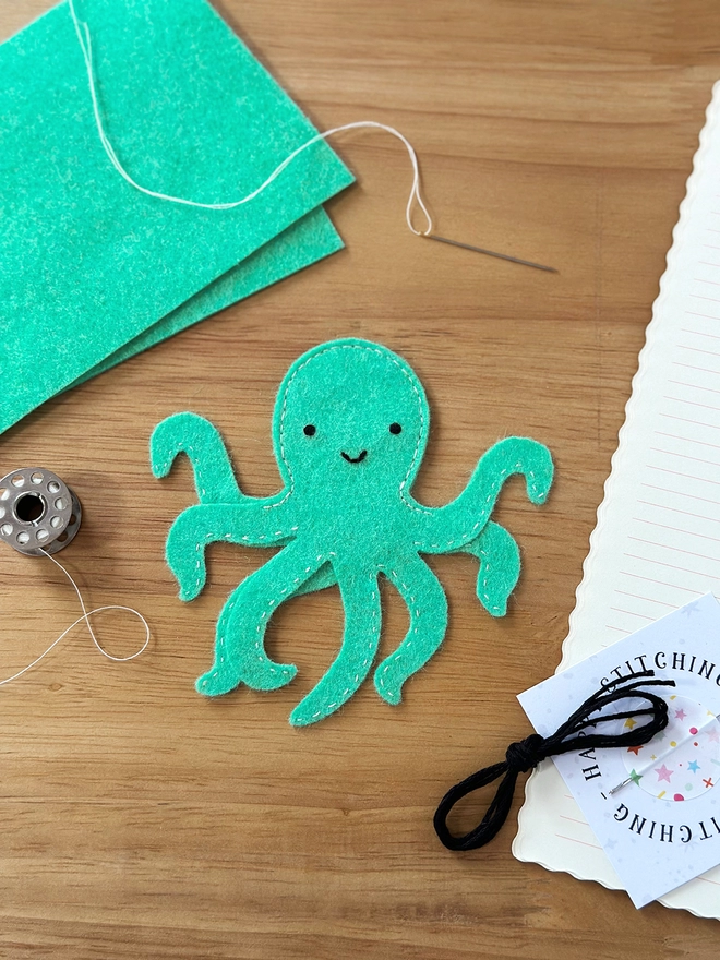 A turquoise felt octopus finger puppet is on a wooden desk beside the craft kit materials that have been used to make it.