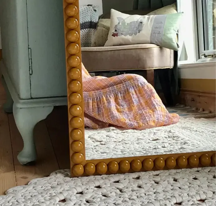 Over-sized bobbin mirror with a turned-bead frame resting on a bedroom floor, leaning against the wall.