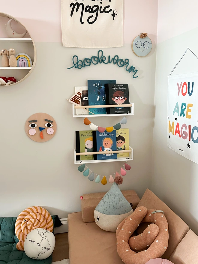 'bookworm' sign for a kids library reading area hanging on the wall above a book shelf filled with modern children's books. Knitted wire word sign handmade by hey kiddo studio