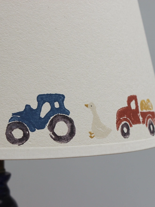 Detailed close-up of Studio Zef's hand-printed farmyard lampshade for children, featuring a navy blue tractor, white goose, red farm truck carrying hay and a pink pig. The colourful countryside illustrations are individually lino printed onto cream card, highlighting the handmade texture and character of this playful children's bedroom lampshade.