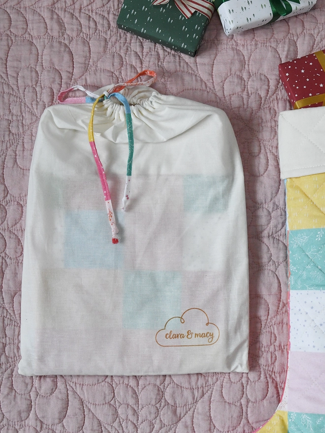 An ivory cotton dust bag lays beside a pastel patchwork stocking on a pink quilt.