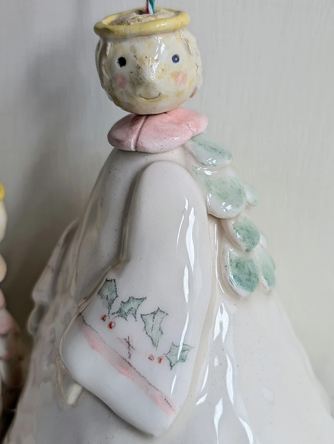 close up of a ceramic angel bell hanging decoration with a pink collar and hand painted holly decoration and green wings 