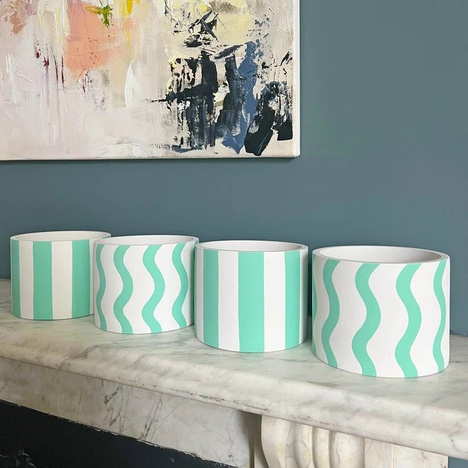 Set of 4 white jesmonite plant pots with mint coloured stripes and squiggles. Set on a fireplace surround with a dark blue background.
