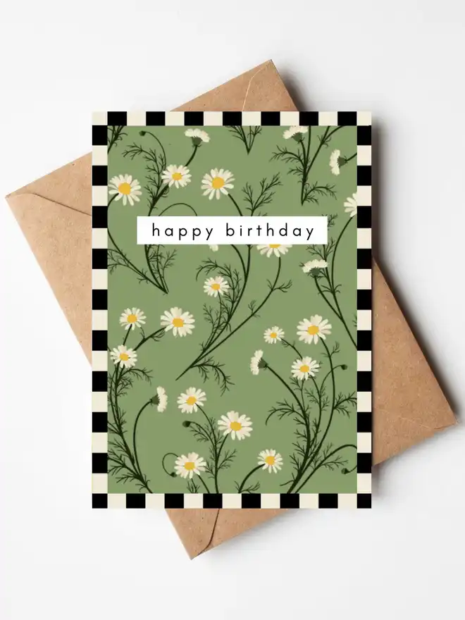 A botanical birthday card featuring hand-illustrated chamomile flowers on a sage green background with a black and cream gingham border, and the words "happy birthday" across the centre. The card is displayed with a kraft envelope.