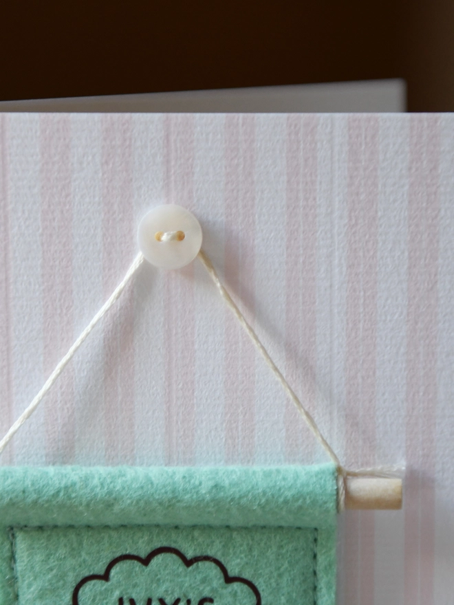 A handmade felt banner is attached to a striped card with a mother of pearl button.