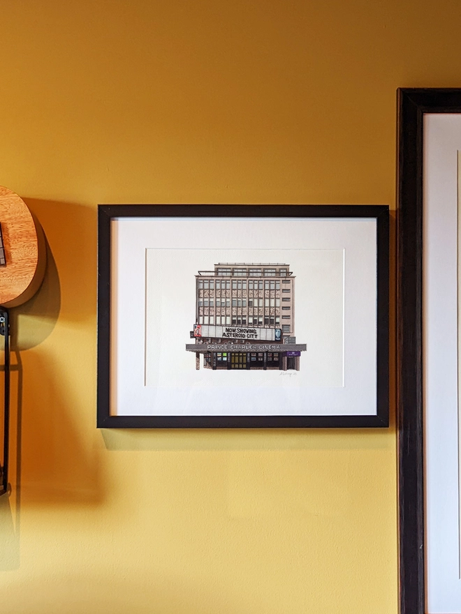 A framed watercolor print of the Prince Charles Cinema hanging on a yellow wall next to a ukulele and another framed artwork.