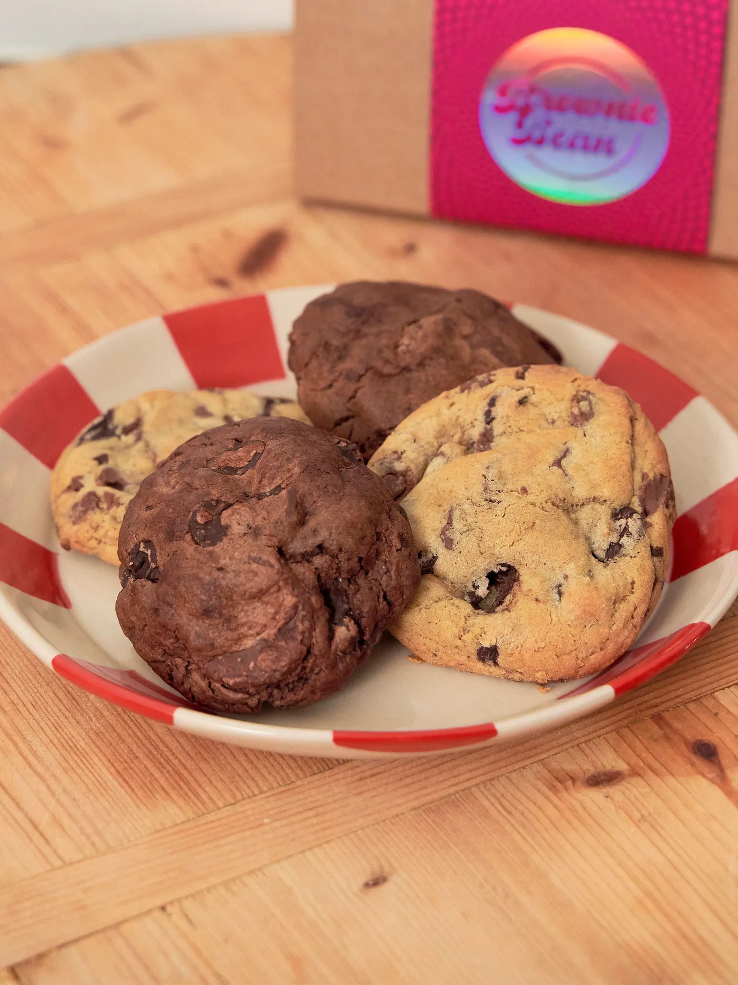 4 chocolate cookies on a red and white plate atop a wooden board with brownie and the bean branded packaging behind