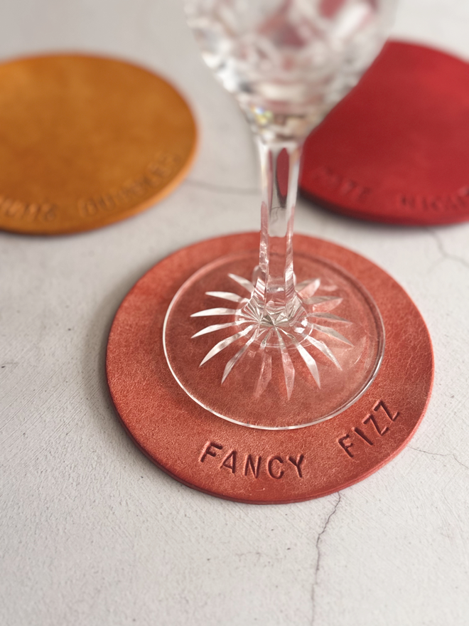pale pink leather coaster with 'fancy fizz' stamped on it, with a champagne glass resting on it. 10cm