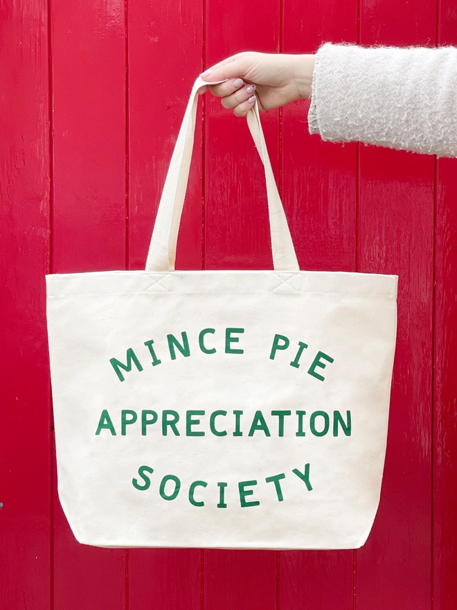 a hand holding out a large natural canvas tote bag bearing the words mince pie appreciation society in front of a red door