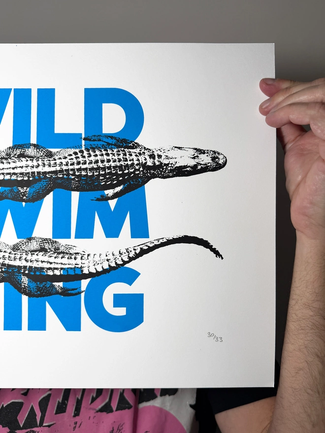 A humorous limited edition A3 screen printed poster. Featuring black alligators over blue 'wild swimming' typography. Showing close up of detail right edge.
