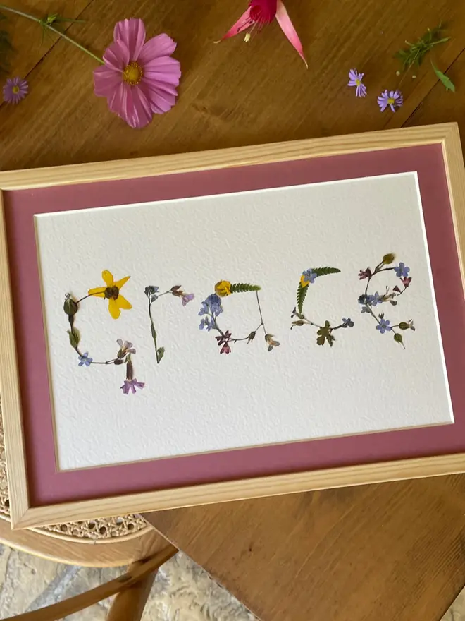 Personalised Original Pressed Flower Name In Frame, a framed flower name prints sitting on a wooden surface. 