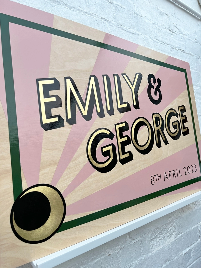 Modern wedding sign featuring the couple's names in gold leaf, outlined in black, and the wedding date, with a sunburst background and a border. Shown against a white brick wall, at an angle. 
