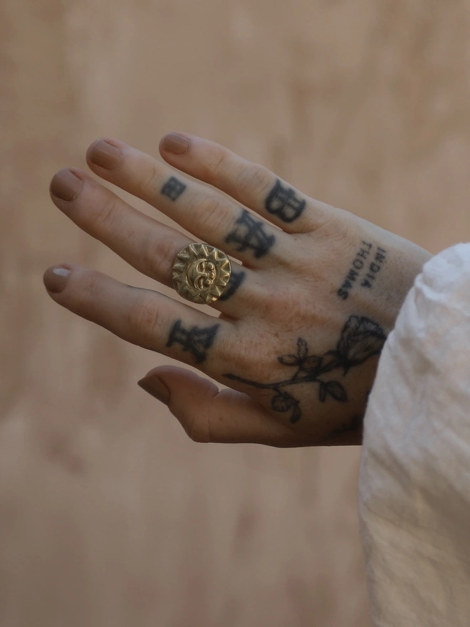 The image of a tattooed female hand wearing a hand carved gold toned brass sun face ring, in front of a rustic terracotta coloured plaster wall
