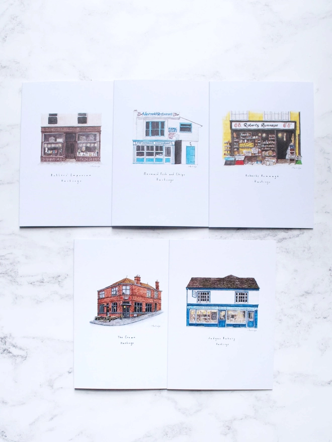 Watercolour illustrated Hastings Old Town shop front cards on A6 white cards. The Crown, Butler's Emporium, Robert's Rummage, Judge's Bakery and Mermaid Fish and Chips are hand painted in watercolour and printed onto cards and displayed layed out in two rows. The background is a pale white marble.