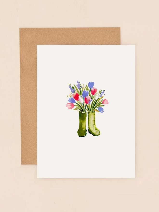 A charming illustration of a pair of green wellington boots filled with a vibrant bouquet of wild spring flowers, including pink tulips, bluebells, and yellow daffodils. The boots are set against a soft, neutral background, highlighting the colorful arrangement. The design exudes a rustic, countryside charm, perfect for conveying warmth and appreciation.