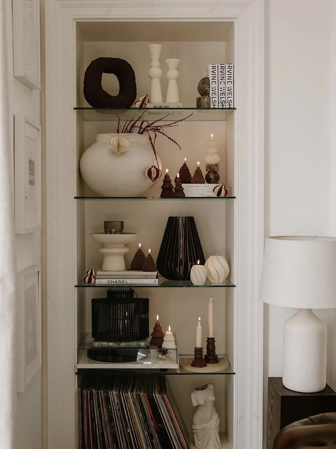 Brown Origami Christmas Tree Warm Candles (Set Of 2). A tall alcove of glass shelves styled with lit brown cone and tree candles, cream sculptural candles, a textured white vase, paper ornaments and a record collection. The earthy candles add a warm, curated glow to a modern living space.