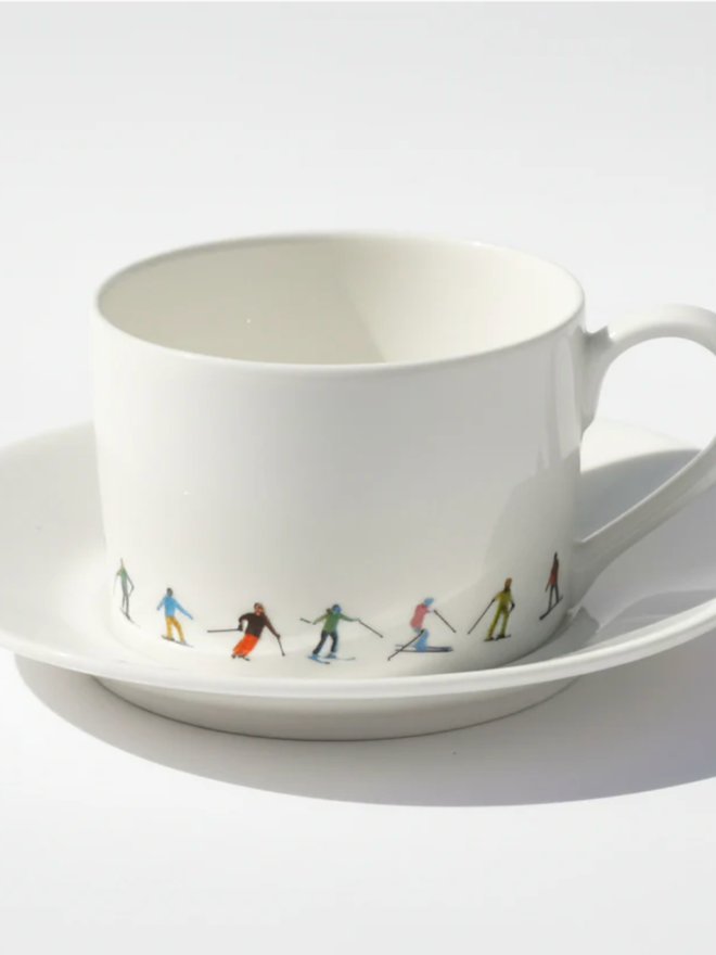 Ski Chain Teacup and Saucer. A teacup and saucer set against a simple white background. The teacup’s lower portion is patterned with a chain of multicoloured skiers.