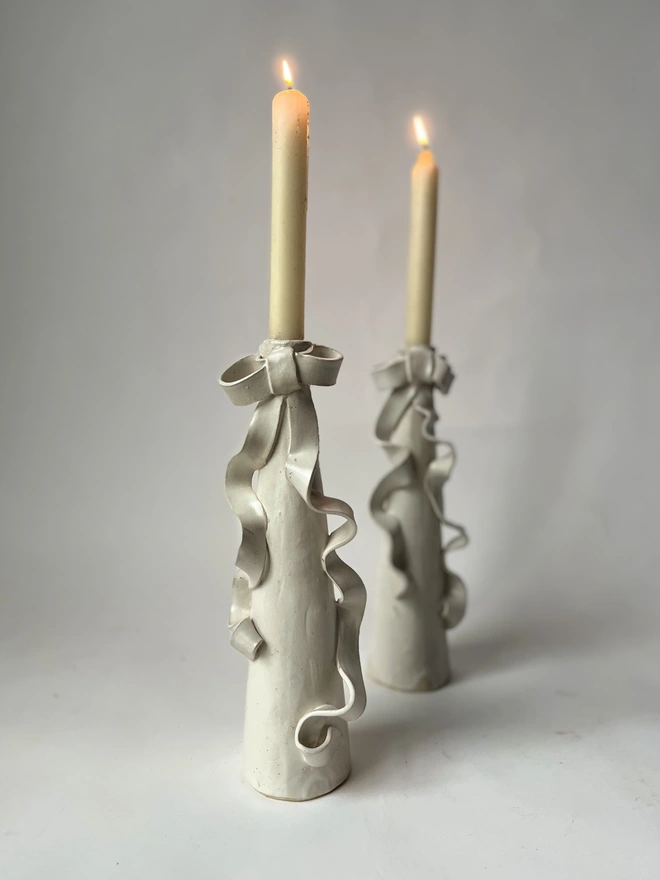 two cream white stoneware tall candlestick holders with a ceramic bow with long ribbons flowing down the body of the candlestick itself, and two lit candles in the holders