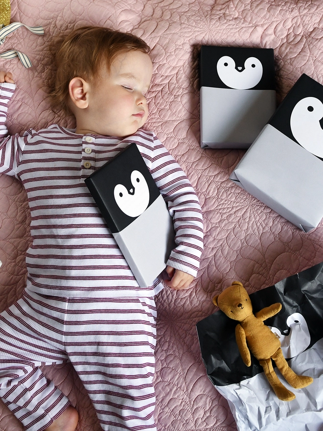 A baby wearing red and white striped pyjamas is asleep on a pink quilt with several gifts around them, all wrapped as penguins.