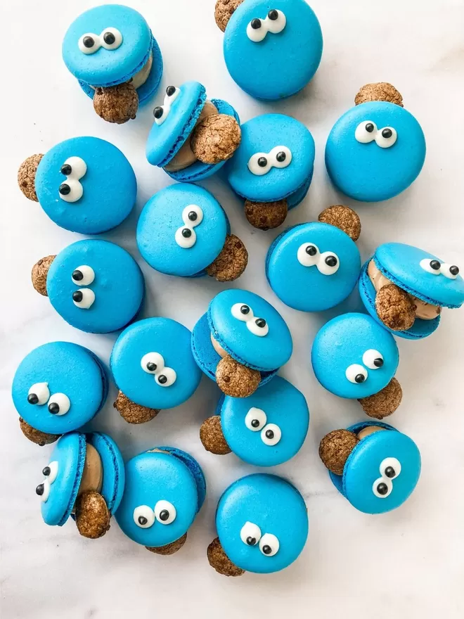blue cookie monster macarons with googly eyes and a chocolate chip cookie in its mouth, sitting in a pile on top of each other