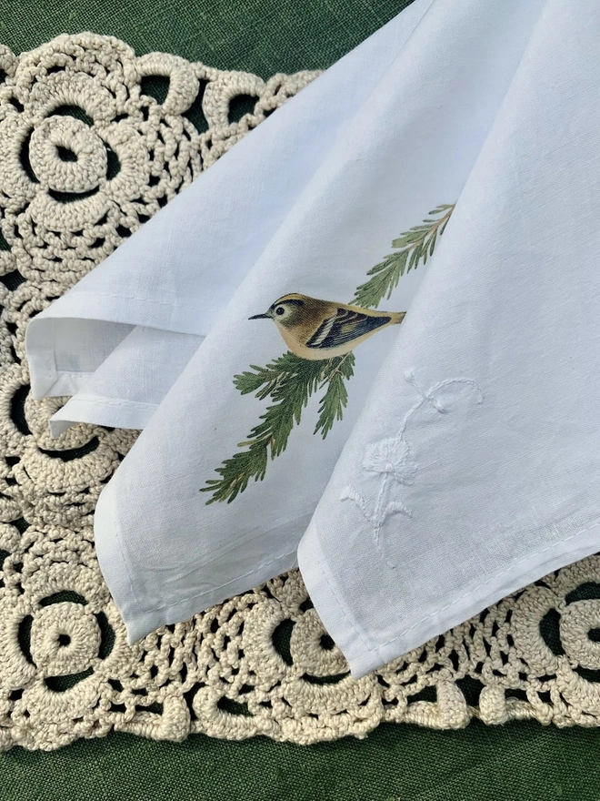A white cotton napkin printed with a vintage illustration of a goldcrest, on a crochet lace table runner