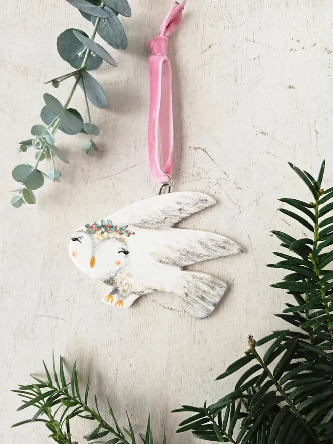 Decorative owl ornament with floral crown hanging from a branch on a soft creme background.