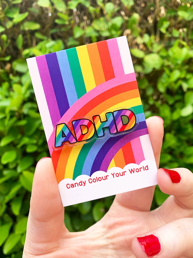 image shows a hand holding a rainbow business card. pinned to the card is an acrylic badge in the shape of the letters ADHD. the letters are filled with diagonal rainbow stripes.