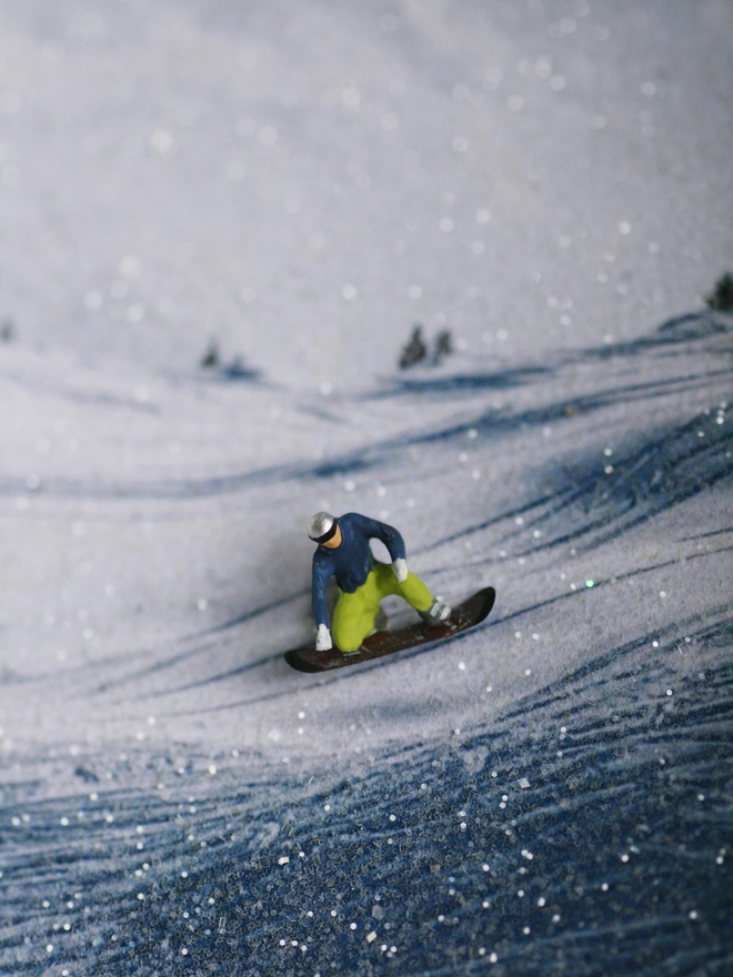 Miniature scene in an artbox showing a tiny male figurine snowboarding against a sparkling mountainous backdrop 