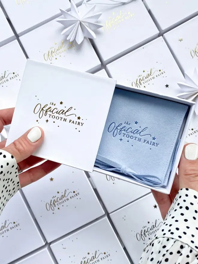 Two hands slide open a gold foil gift box to show the blue embossed pouch inside. Stacks of matching boxes fill the background.