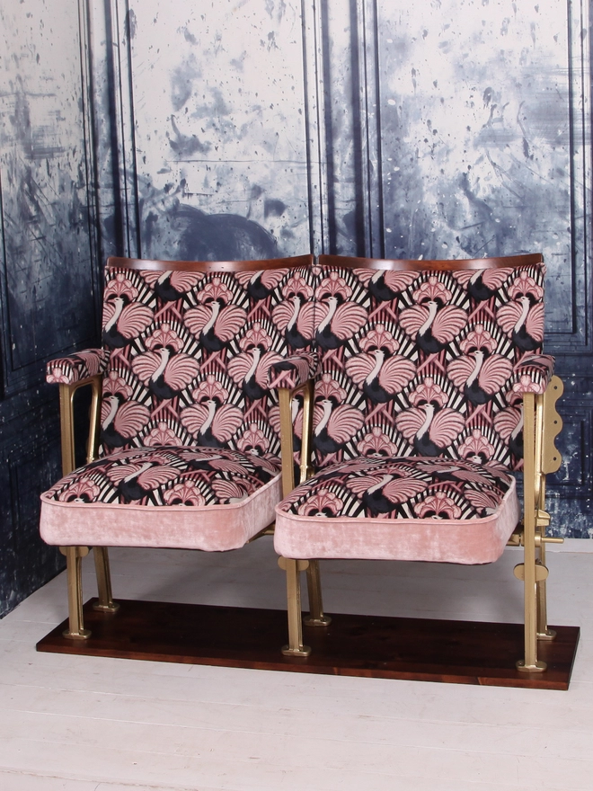 Set of two vintage cinema seats upholstered in a pink ostrich Art Deco design velvet with pink panel and piping against a blue marbled wall.  Seats are down